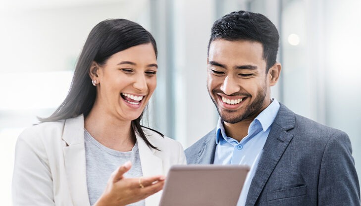 Eine Frau und ein Mann stehen zusammen und betrachten lachend einen Tablet Bildschirm.