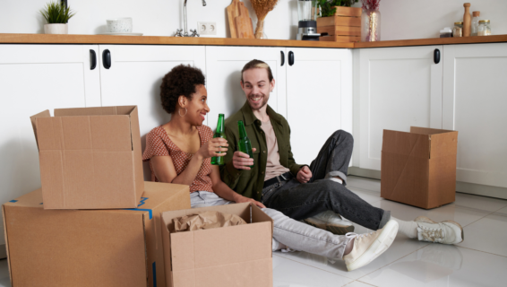Zwei Personen sitzen in einer Wohnung inmitten von Kartons auf dem Boden und stoßen mit ihren Bierflaschen an