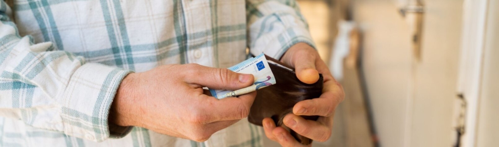 Mann mit kariertem Hemd hält sein Portemonnaie in der Hand mit einem 20 Euro Schein