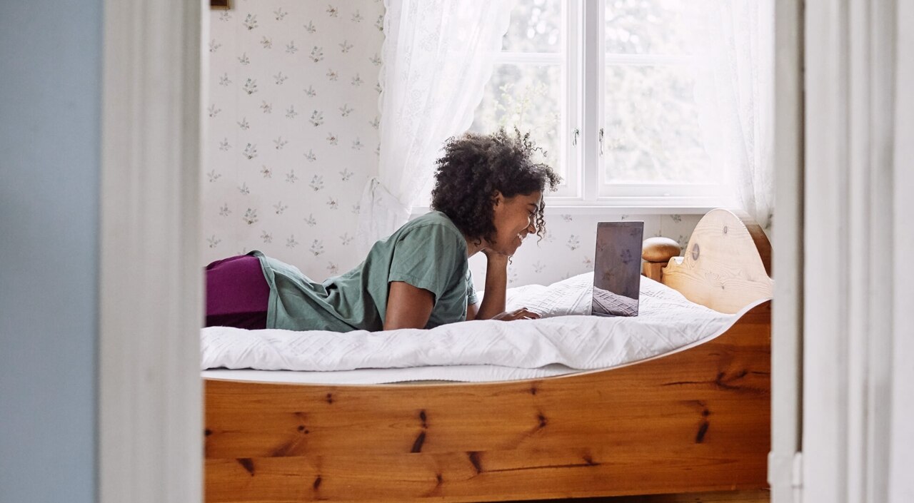 Junge Frau liegt mit ihrem Laptop auf dem Bett in einem hellen, freundlichen Zimmer