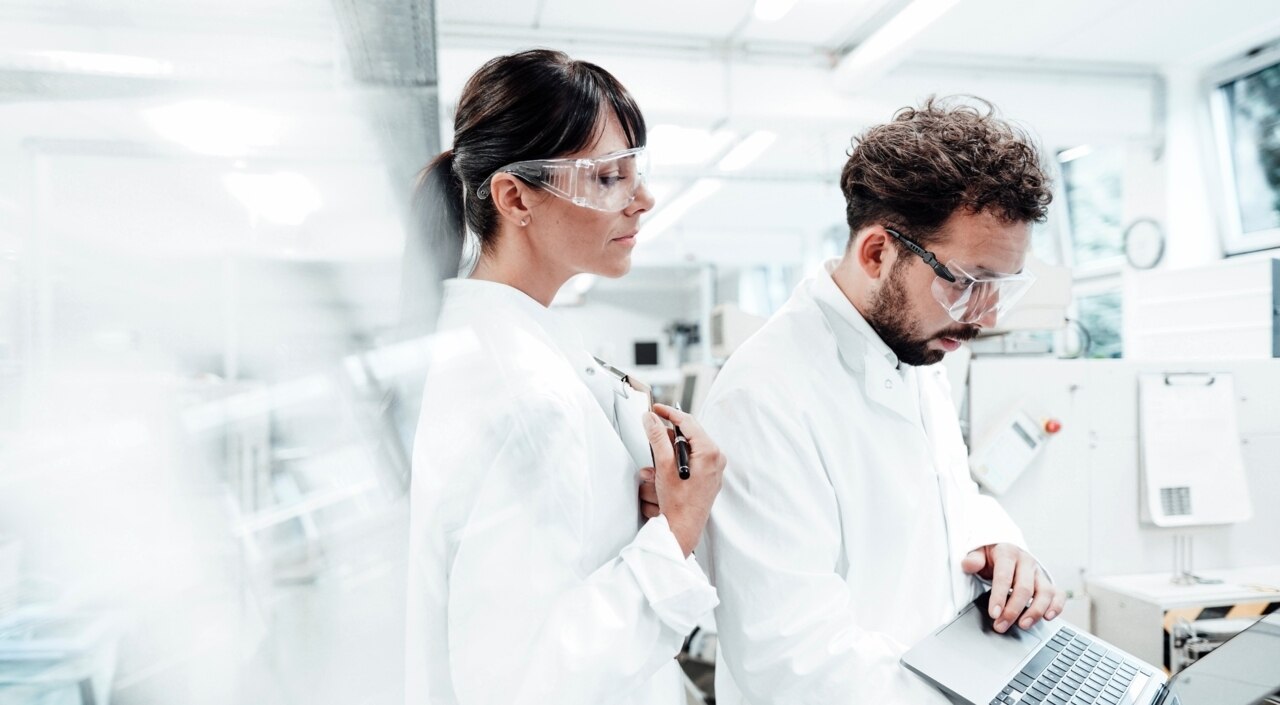 Eine Frau und ein Mann mit weißem Kittel und Schutzbrille stehen in einem Labor und schauen zusammen auf einen Laptop