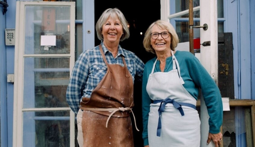 Zwei ältere Arbeitnehmerinnen stehen lächelnd vor einer Werkstatt.