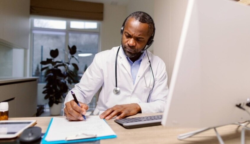 Ein Arzt im weißen Kittel sitzt am Schreibtisch, hat ein Headset auf und schreibt etwas auf.