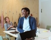 Lächelnde Kolleg*innen sitzen mit Laptops am Schreibtisch.