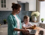 Berufstätige Mutter in der Küche am Laptop, daneben ihr frühstückendes Kind: Staatliche Hilfen für Familien ermöglichen die Vereinbarkeit von Beruf und Elternschaft.