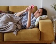 Junge brünette Frau liegt unter einer Decke auf dem Sofa und scrollt auf ihrem Smartphone