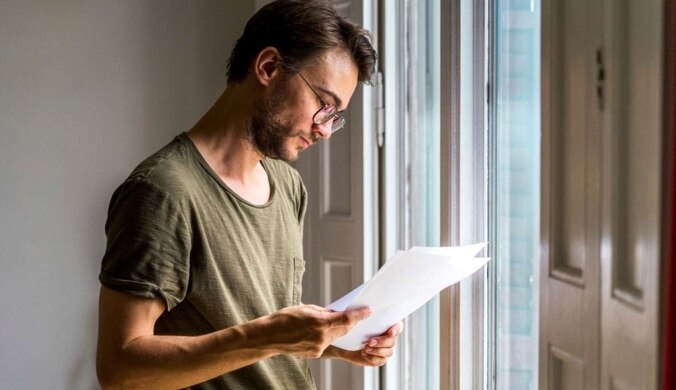 Junger Mann steht am Fenster und sieht sich einige Papierseiten an.