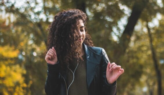 Junge Frau in Lederjacke und mit Kopfhörern tanzt mit geschlossenen Augen durch den Park.