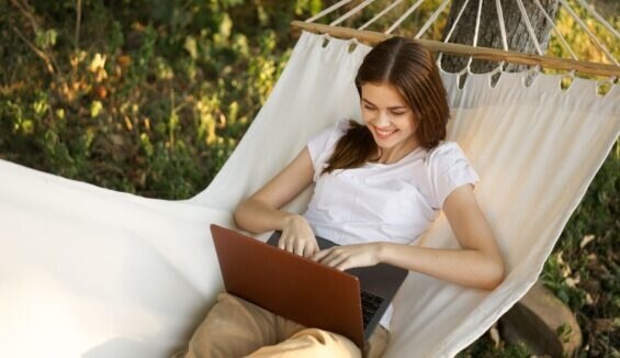 Eine Frau sitzt in einer Hängematte und schaut lächelnd auf einen Laptop.
