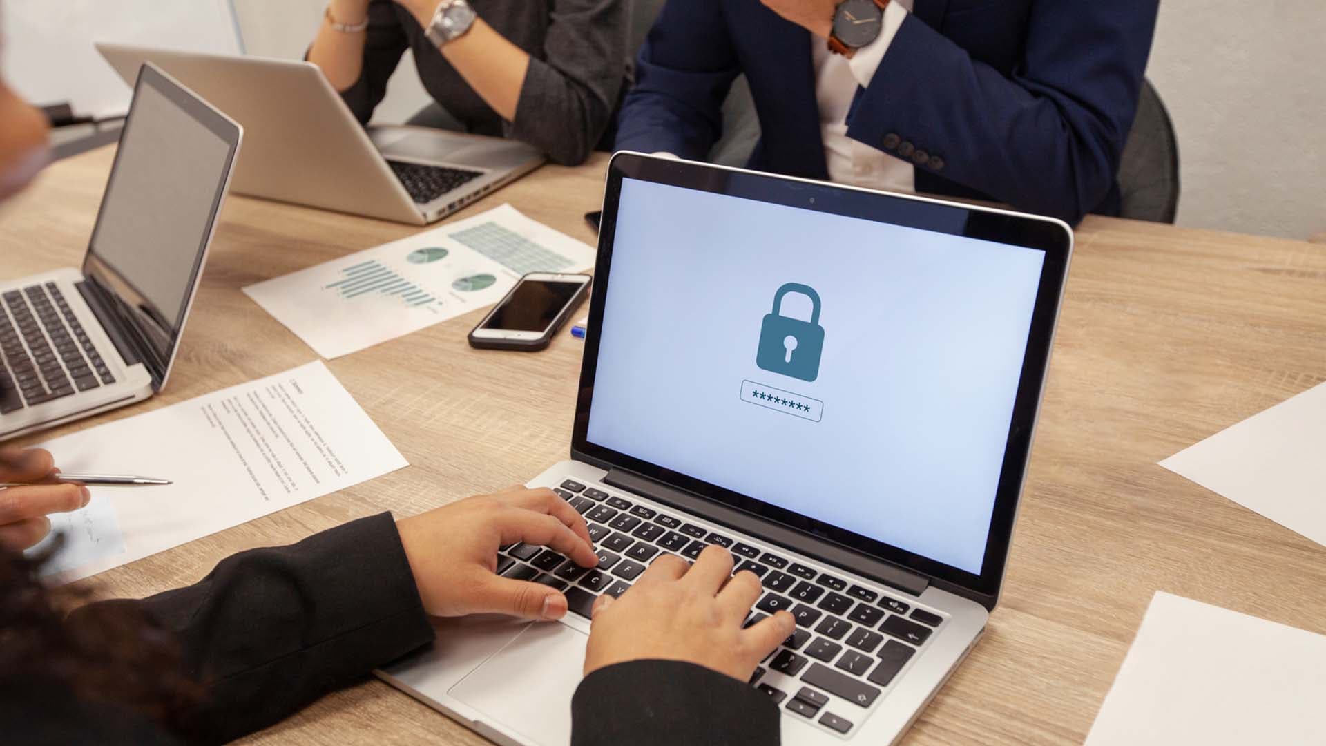 Nahaufnahme eines Mitarbeitenden, der mit anderen Mitarbeitenden am Tisch sitzt und sein Passwort in einen Laptop eingibt.