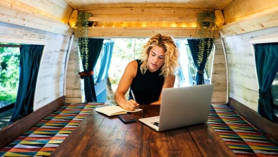 Eine Frau sitzt in einem Wohnwagen an einem Tisch. Ihr Laptop steht vor ihr und sie schreibt etwas in ihr Notizbuch.
