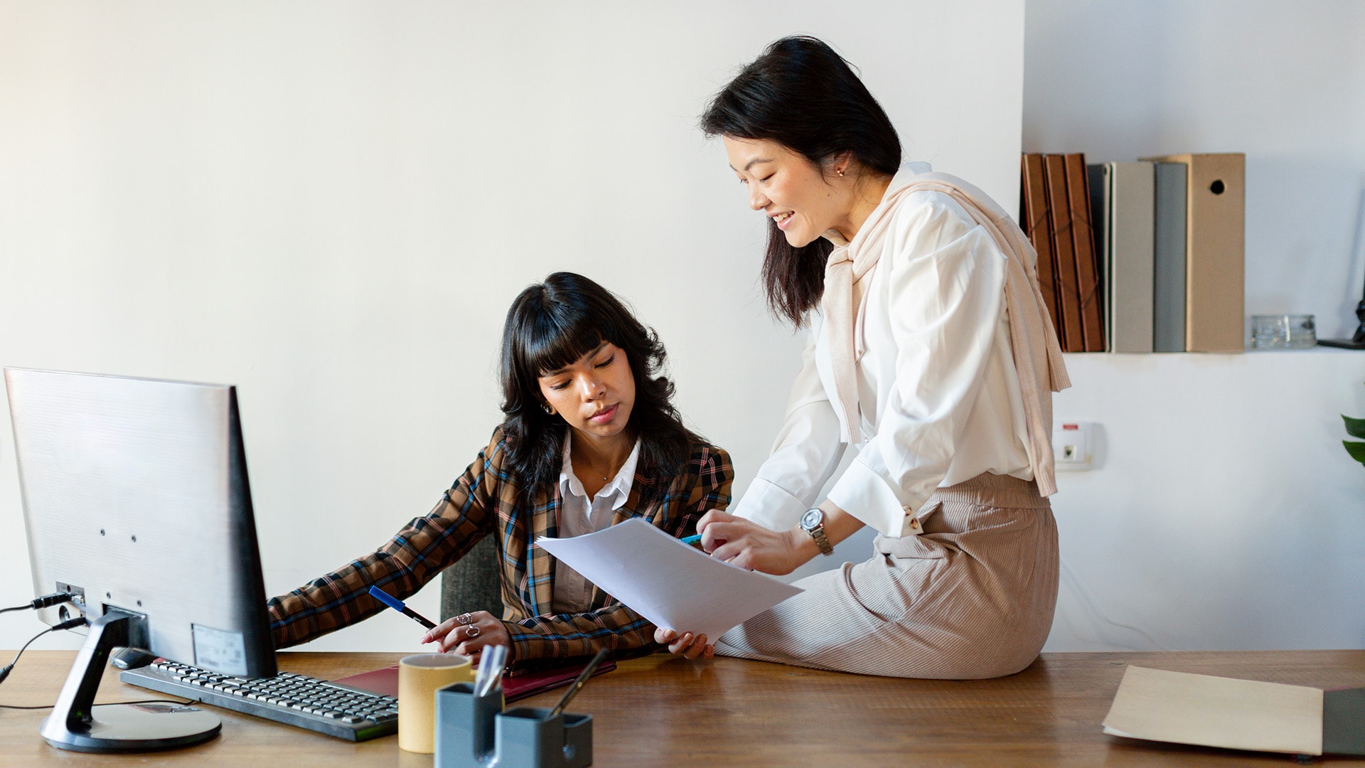 Eine elegante Frau asiatischer Herkunft sitzt auf dem Schreibtisch ihrer Kollegin und zeigt ihr ein Blatt, während ihre Kollegin auf ihrem Computer tippt.