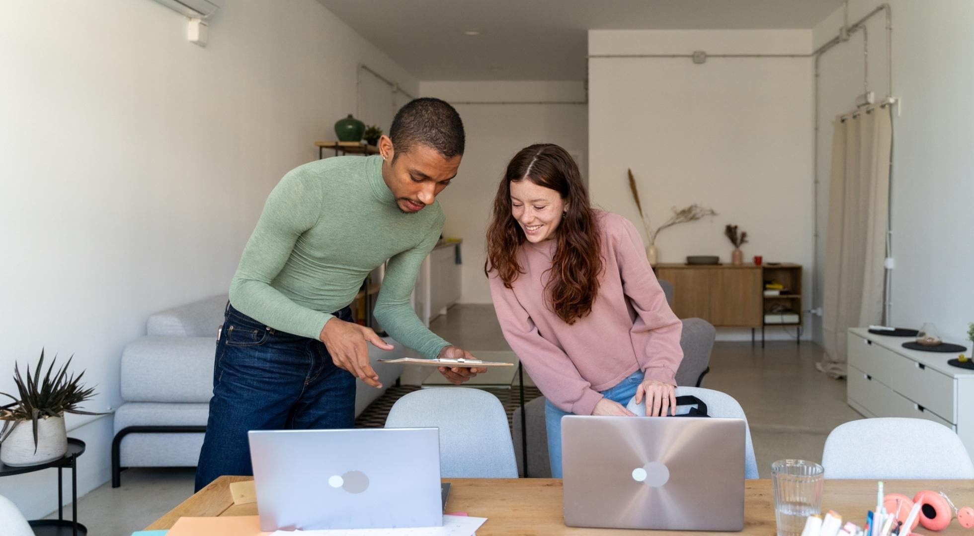 Zwei junge Menschen stehen an einem Tisch, auf dem zwei Laptops stehen. Der Mann zeigt der Frau etwas auf einem Klemmbrett.