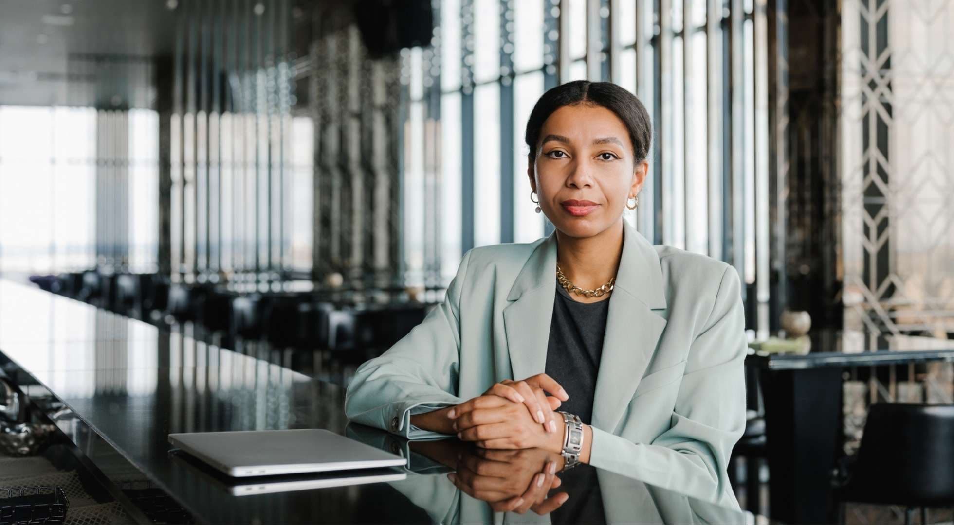 Eine Frau in Business-Kleidung sitzt an einem langen Tisch und schaut ernst in die Kamera. Vor ihr steht ein geschlossener Laptop.