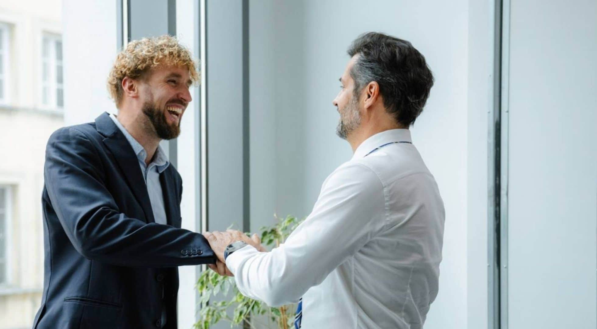 Zwei Männer stehen sich im Büro gegenüber, geben sich die Hand und lachen.