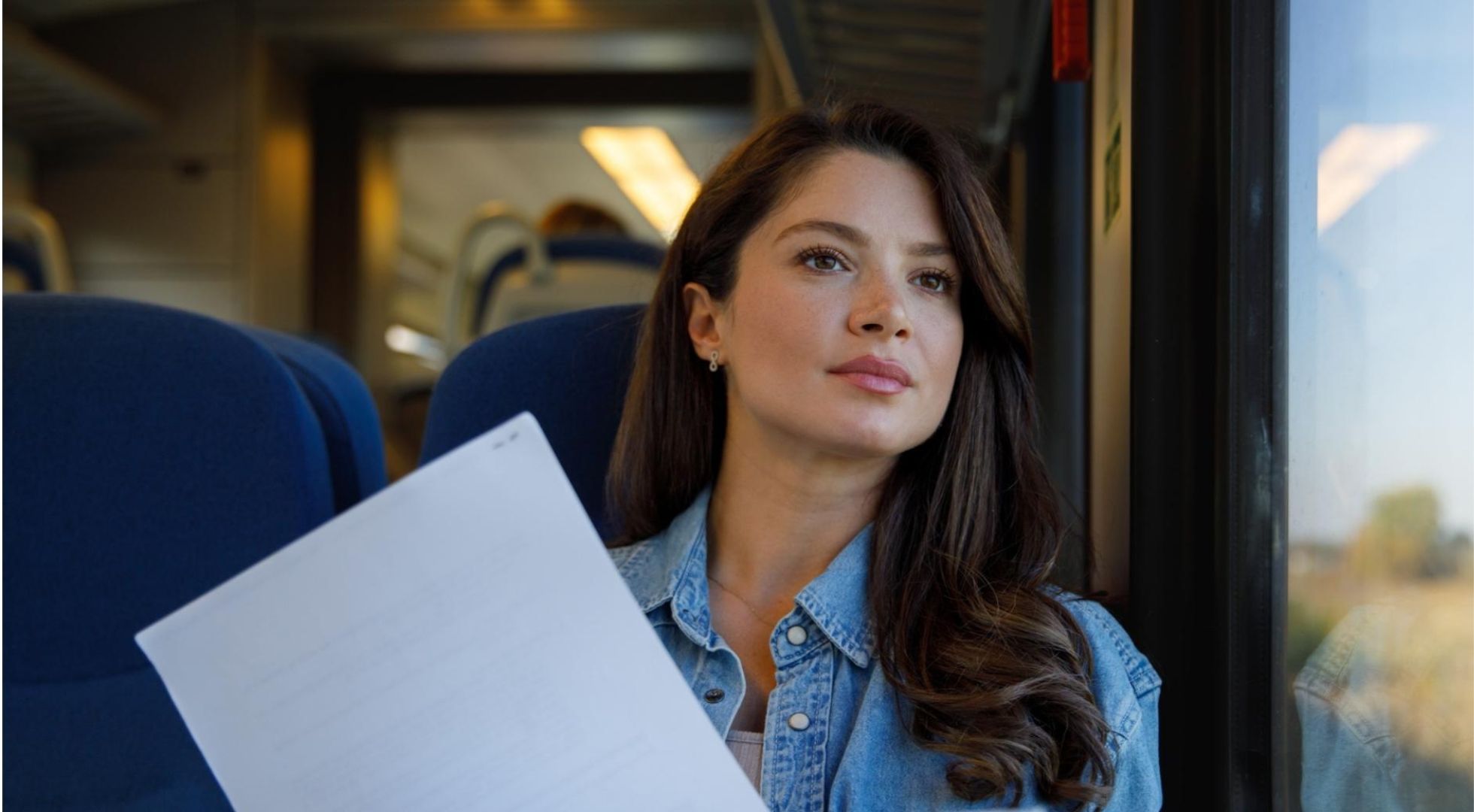 Eine Frau sitzt in. der Bahn und schaut aus dem Fenster. Sie hält Zettel in der Hand.