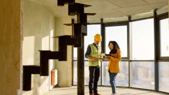 Ein Architekt mit einem gelben Helm steht in einer Doppelhauswohnung mit großen transparenten Fenstern und unterhält sich mit einer Frau in Blazer