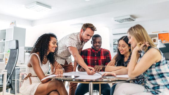 Eine gemischte Gruppe von fünf Kollegen unterschiedlicher Herkunft sitzt in einem hellen Büro um einen Tisch herum: Auf dem Tisch sind Blätter ausgebreitet und einer der Kollegen zeigt mit einem Bleistift auf ein Blatt.
