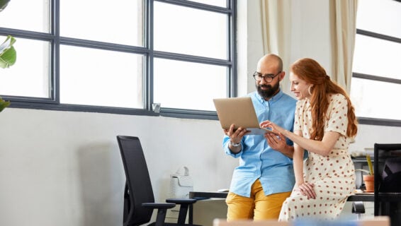 Zwei Kollegen, ein kahlköpfiger Mann mit Bart und eine rothaarige Frau mit langen Haaren, sitzen mit einem Laptop in der Hand auf der Kante eines Schreibtisches.