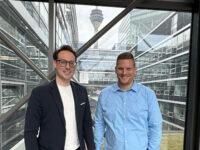 Christoph Schulz, EMEA Talent Acquisition Manager bei Orion Carbons, und Martin Spielvogel, Head of Recruiting Germany & Switzerland bei Henkel, stehen vor einer Glasscheibe im Flur des Stepstone Gebäudes. Hinter ihnen sieht man einen Innenhof mit Gras. In der Ferne sieht man die Spitze des Düsseldorfer Turms.