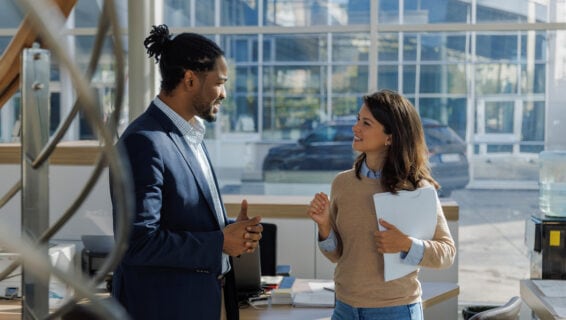 Eine Personalvermittlerin steht in einem verglasten Büro. Sie hält Unterlagen in der Hand und unterhält sich mit einem jungen Bewerber in Hemd und Blazer.