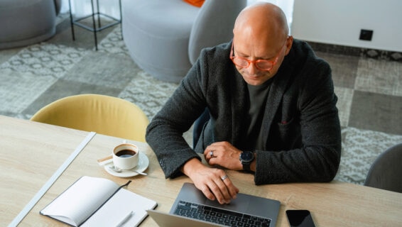 Ein kahlköpfiger Mann mit einer orangefarbenen runden Brille in einem eleganten schwarzen Wollmantel sitzt mit seinem Laptop, einem Kaffee und einem Notizbuch an einem Tisch.