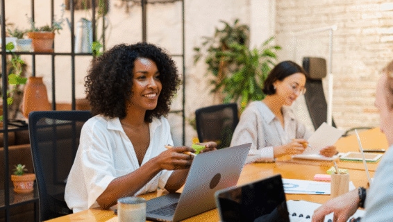 Eine junge Frau mit lockigem braunem Haar unterhält sich in einem Großraumbüro mit einem Kollegen. Das Büro ist hell und mit Pflanzen ausgestattet, was eine gemütliche Atmosphäre schafft.