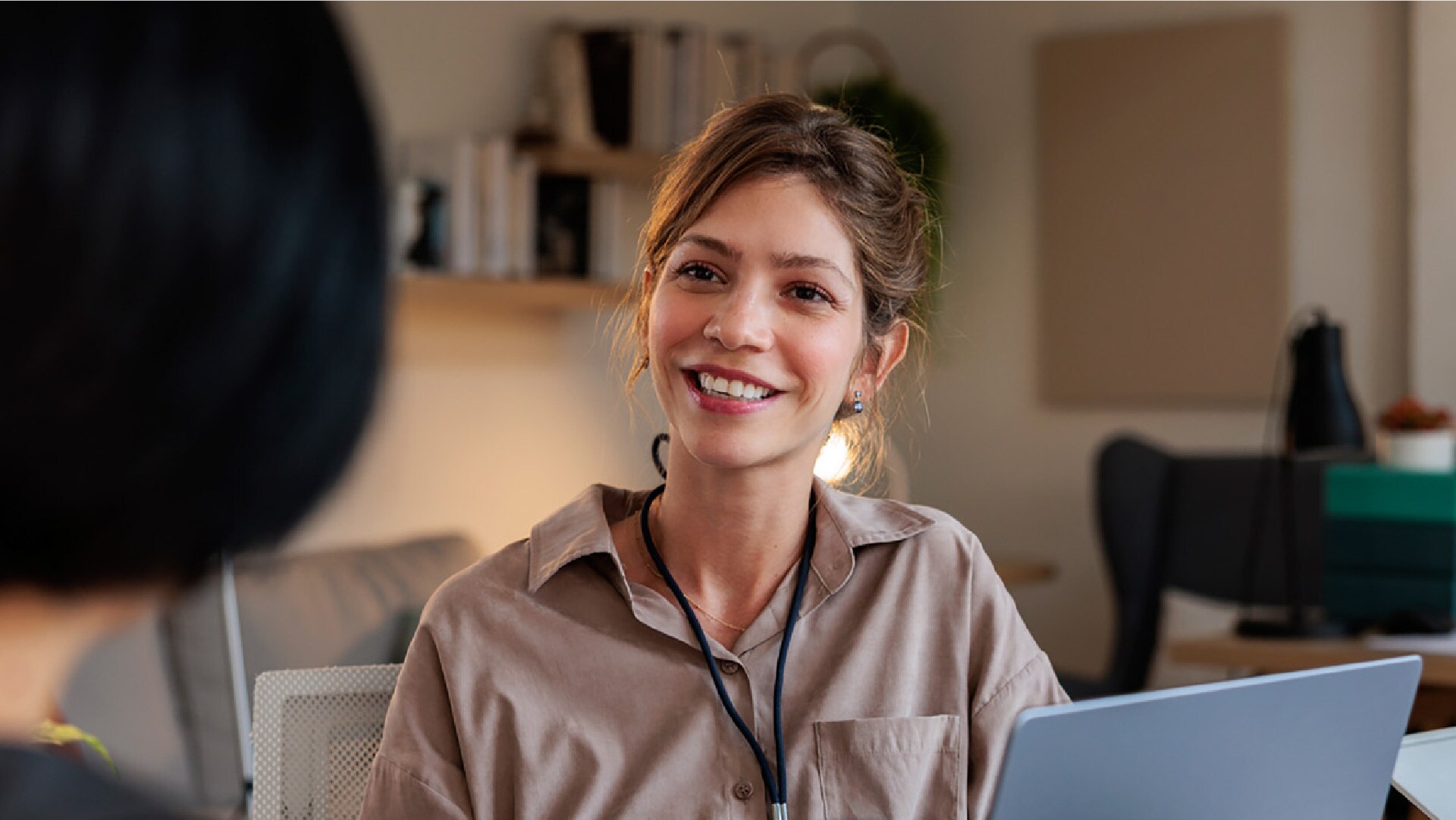 Eine Personalvermittlerin sitzt vor ihrem Laptop und lächelt eine Bewerberin an, die man von hinten sieht.