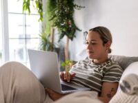 Junge Frau liegt auf der Couch und arbeitet am Laptop.